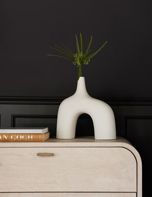 The Leonor larger sculptural arched matte white ceramic Vase sitting atop a white washed dresser next to a stack of books in front of a black accented wall