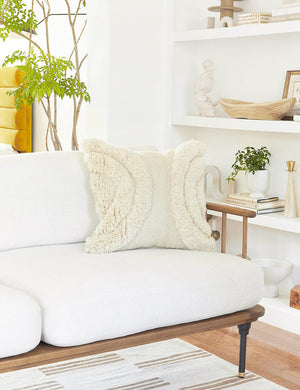 The arches ivory square pillow sits atop a white linen sofa with a wooden frame with a decor filled shelf in the background