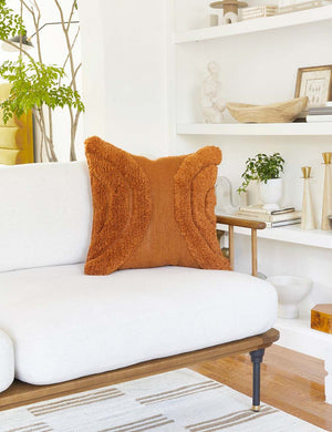 The arches rust square pillow sits atop a white linen sofa with a wooden frame with a decor filled shelf in the background