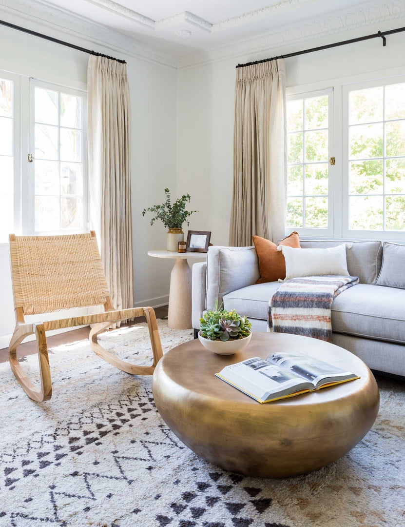 | The Arteriors bates round metallic coffee table sits in a living room with a woven accent chair, a gray linen sofa, and a geometric patterned rug.