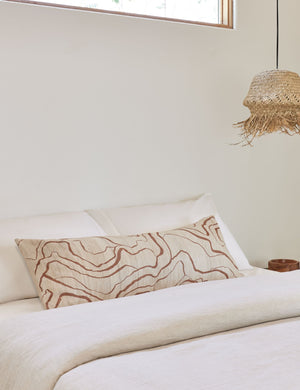 Canyon Terracotta Long Lumbar Pillow lays on a bed with white linens next to a jute pendant light