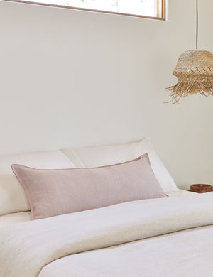 The arlo Greige long lumbar pillow lays on a bed with ivory linens with a jute pendant light hanging next to it
