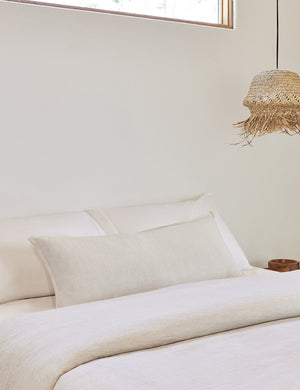 The arlo Ivory long lumbar pillow lays on a bed with ivory linens with a jute pendant light hanging next to it