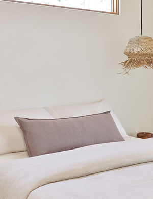 The arlo Dark Natural long lumbar pillow lays on a bed with ivory linens with a jute pendant light hanging next to it