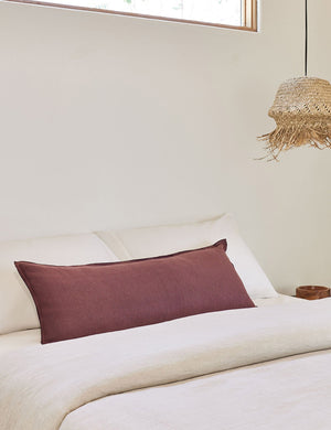 The arlo Aubergine burgundy long lumbar pillow lays on a bed with ivory linens with a jute pendant light hanging next to it
