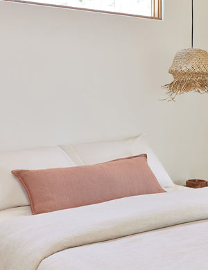 The arlo Terracotta long lumbar pillow lays on a bed with ivory linens with a jute pendant light hanging next to it