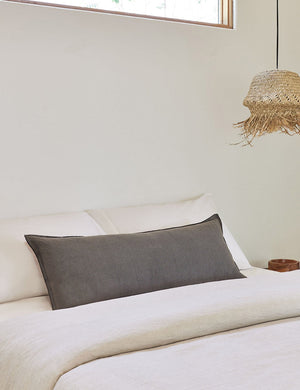 The arlo Conifer gray long lumbar pillow lays on a bed with Ivory linens with a jute pendant light hanging next to it