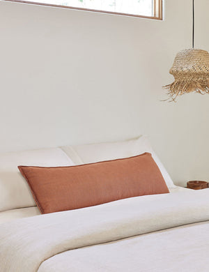 The arlo rust orange long lumbar pillow lays on a bed with ivory linens with a jute pendant light hanging next to it