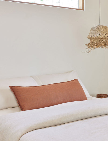 #color::rust #style::long-lumbar | The arlo rust orange long lumbar pillow lays on a bed with ivory linens with a jute pendant light hanging next to it