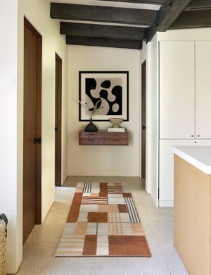 The Benita natural toned hand-knotted floor rug by Nina Freudenberger in its runner size lays in a hallway with a wooden shelf, a black sculptural vase, and an abstract wall art