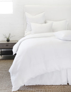 The white blair stonewashed linen duvet by pom pom at home lays atop a white linen framed bed in a bedroom with a jute area rug, a white brick wall, and a wooden nightstand