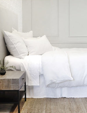 The Blair white linen euro, king, and standard shams by pom pom at home laying on a white linen framed bed in a bedroom with a white brick wall, a jute rug, and a wooden nightstand