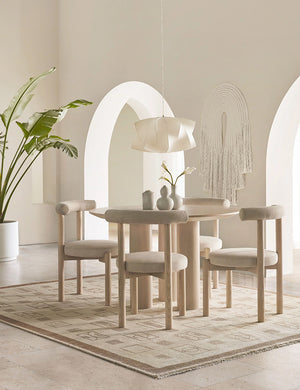 Mojave Round Dining Table sits in a cream toned room surrounded by cream cushioned dining chairs atop a cream and neutral patterned rug