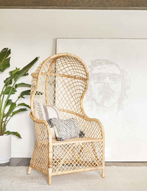 The Charley rattan accent chair with sloping back sits in a bright room with a beige rug, white vase, and large portrait wall art in the background