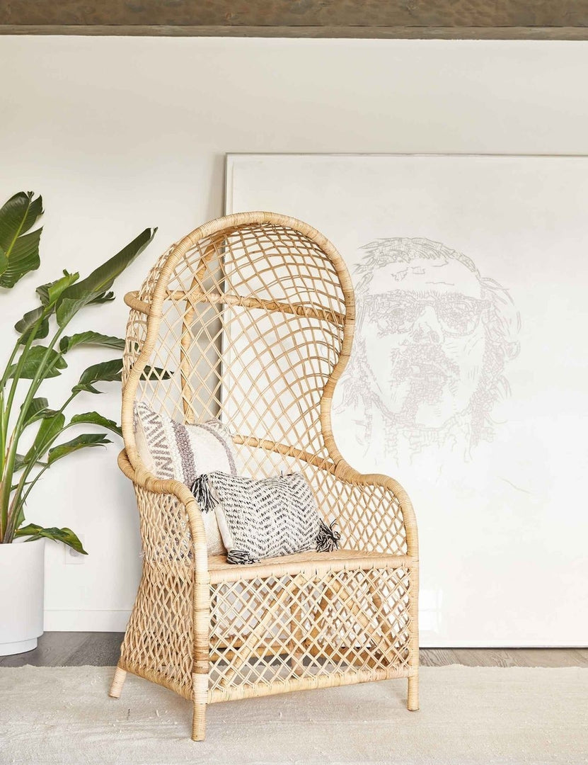 | The Charley rattan accent chair with sloping back sits in a bright room with a beige rug, white vase, and large portrait wall art in the background