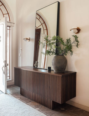 The Evangeline diffused white glass orb sconces with brass frames sit in an entryway with a striped dark wooden sideboard and a tall rectangular mirror between them