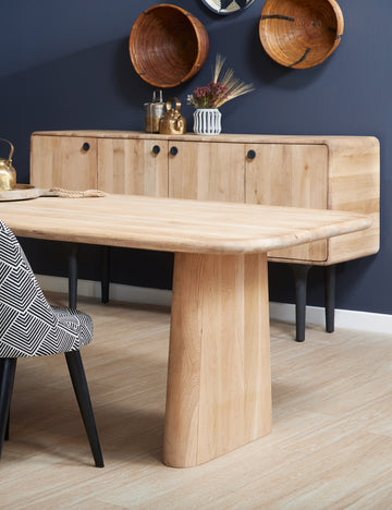 #color::natural | The Mela oak wood dining table sits in a dining room with a white and black patterned chair and an oak wood side table with four cabinets below wooden wall hangings.