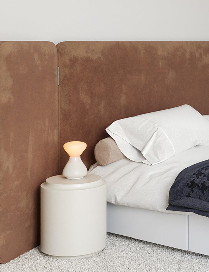 | The Reflection Noma white table lamp by Tala sits in a bedroom atop a white nightstand with a brown velvet framed bed to its right