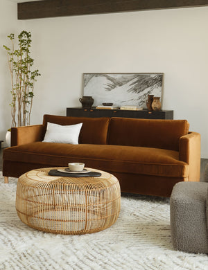 Cognac Velvet Belmont Sofa sits in a living room with a round woven coffee table, a plush brown and white rug, and a black sideboard