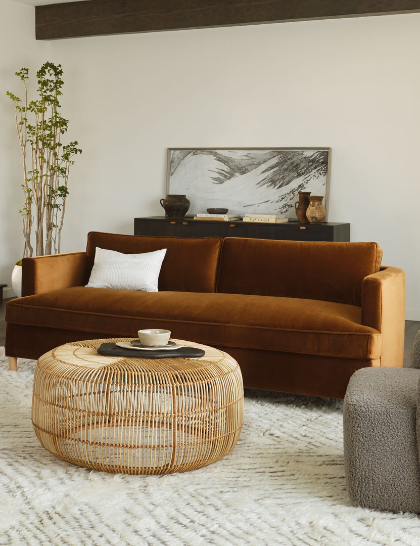 #size::72-W #size:84-W #color::cognac #size::96-W | Cognac Velvet Belmont Sofa sits in a living room with a round woven coffee table, a plush brown and white rug, and a black sideboard
