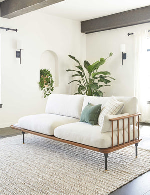 The Estie white rug with thick braids sits in a bright living room with a cushioned wooden sofa, black beamed ceilings, and plants throughout the room.