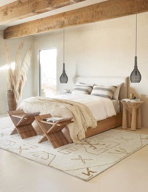 The Evet neutral geometric wool floor rug lays in a neutral bedroom with an upholstered bed with wooden bedrails and two hanging black pendant lights on either side of the bed.