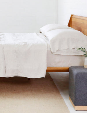 The flax Linen Sheet Set by Pom Pom at Home lays on a wooden framed bed in a bedroom with a gray nightstand and a jute rug