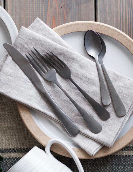 | The Woodstock stainless steel 5-piece flatware set by farmhouse pottery sits on a dining table atop a ceramic plate and beige linen napkins