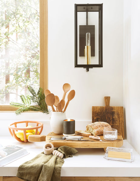 #color::terracotta | The Prong terracotta ceramic centerpiece bowl by SIN sits on a white countertop in a kitchen with wooden cutting and cheese boards, olive green linens, and a black sconce light with golden hardware
