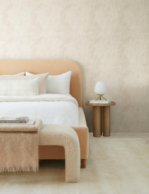 The Delta natural wooden side table with pedestal base sits in a bedroom with a peach velvet bed, a white boucle bench, and gray patterned walls