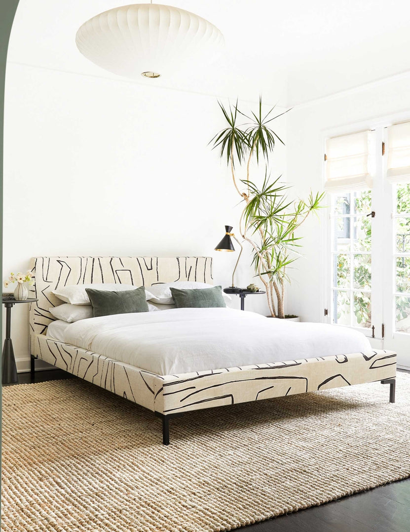 #color::grafitto #size::twin #size::full #size::queen #size::king #size::cal-king | The Deva Grafitto platform bed lays in a bright bedroom in between two round side tables and atop a jute rug