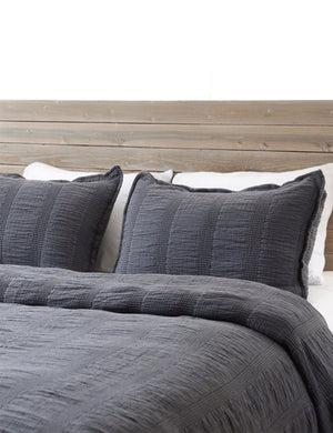 The Nantucket Cotton Matelassé midnight gray Sham by Pom Pom at Home sits on a wooden framed bed with other gray Matelassé linens