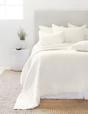The Hampton cream Quilted Coverlet by Pom Pom at Home laying on a white linen framed bed in a bedroom with a jute rug and a white brick wall