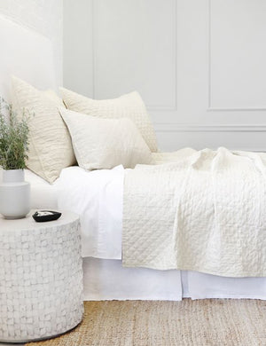 Side view of the Hampton cream Quilted Coverlet by Pom Pom at Home laying on a white linen framed bed in a bedroom with a jute rug and a white brick wall