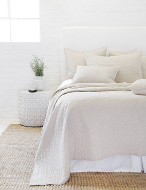 Hampton flax Quilted Coverlet by Pom Pom at Home lays on a white linen framed bed in a bedroom with a jute rug and a white brick wall