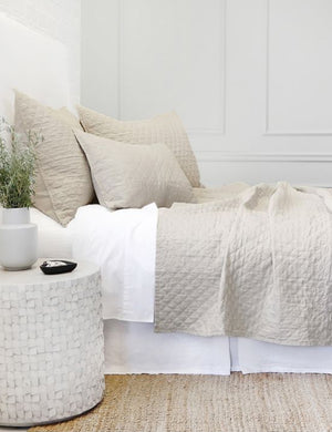 Side view of the Hampton flax Quilted Coverlet by Pom Pom at Home laying on a white linen framed bed in a bedroom with a jute rug and a white brick wall