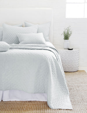 The Hampton light blue Quilted Coverlet by Pom Pom at Home lays on a white linen framed bed in a bedroom with a jute rug and a white brick wall