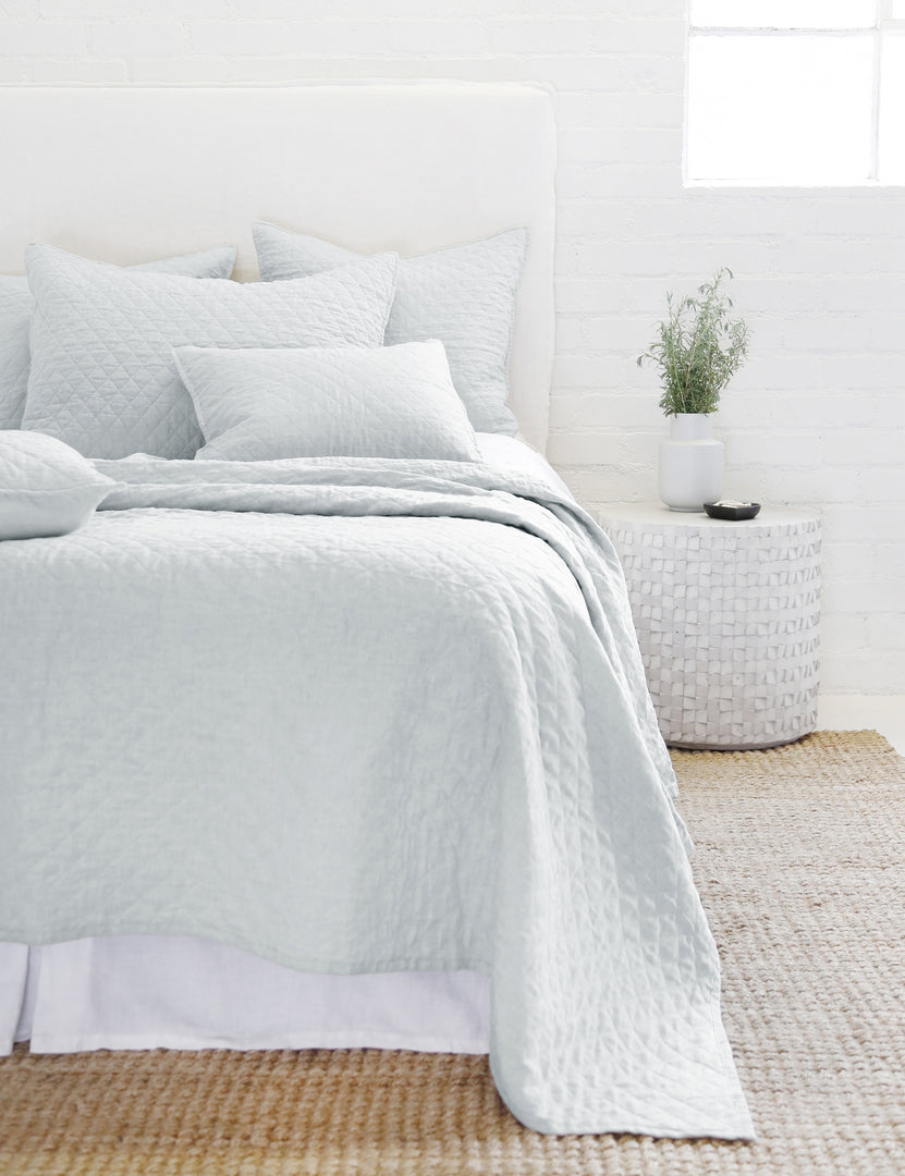 #color::ocean #size::king #size::queen #size::twin | The Hampton light blue Quilted Coverlet by Pom Pom at Home lays on a white linen framed bed in a bedroom with a jute rug and a white brick wall