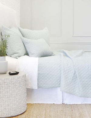 Side view of the Hampton light blue Quilted Coverlet by Pom Pom at Home laying on a white linen framed bed in a bedroom with a jute rug and a white brick wall