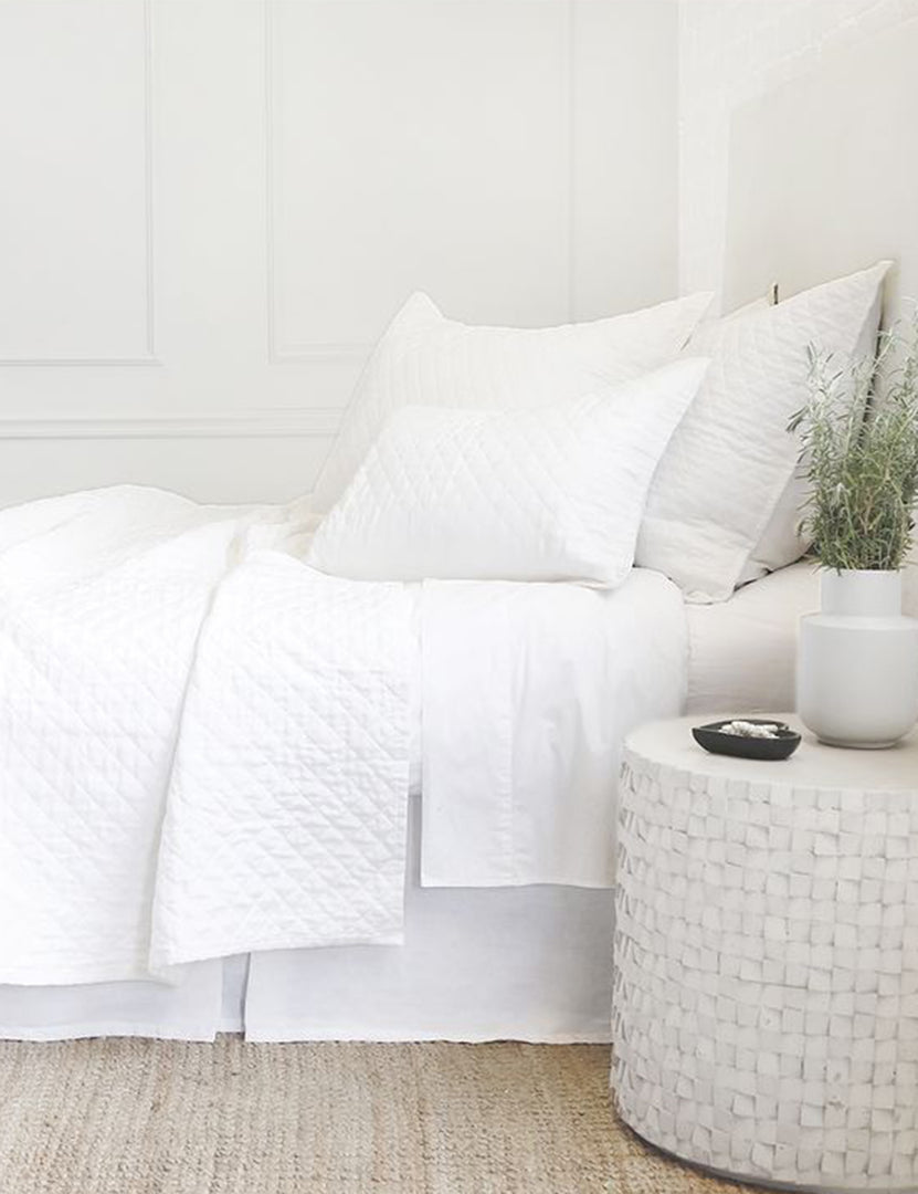 #color::white #size::king #size::queen #size::twin | Side view of the Hampton white Quilted Coverlet by Pom Pom at Home laying on a white linen framed bed in a bedroom with a jute rug and a white brick wall