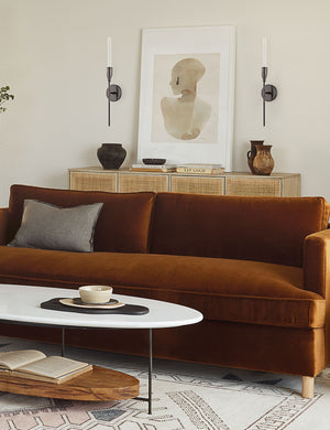 Two Kama sleek, industrial black sconces with an aged bronze finish sits in a living room with a portrait painting in between them, a cognac velvet sofa, and a woven sideboard