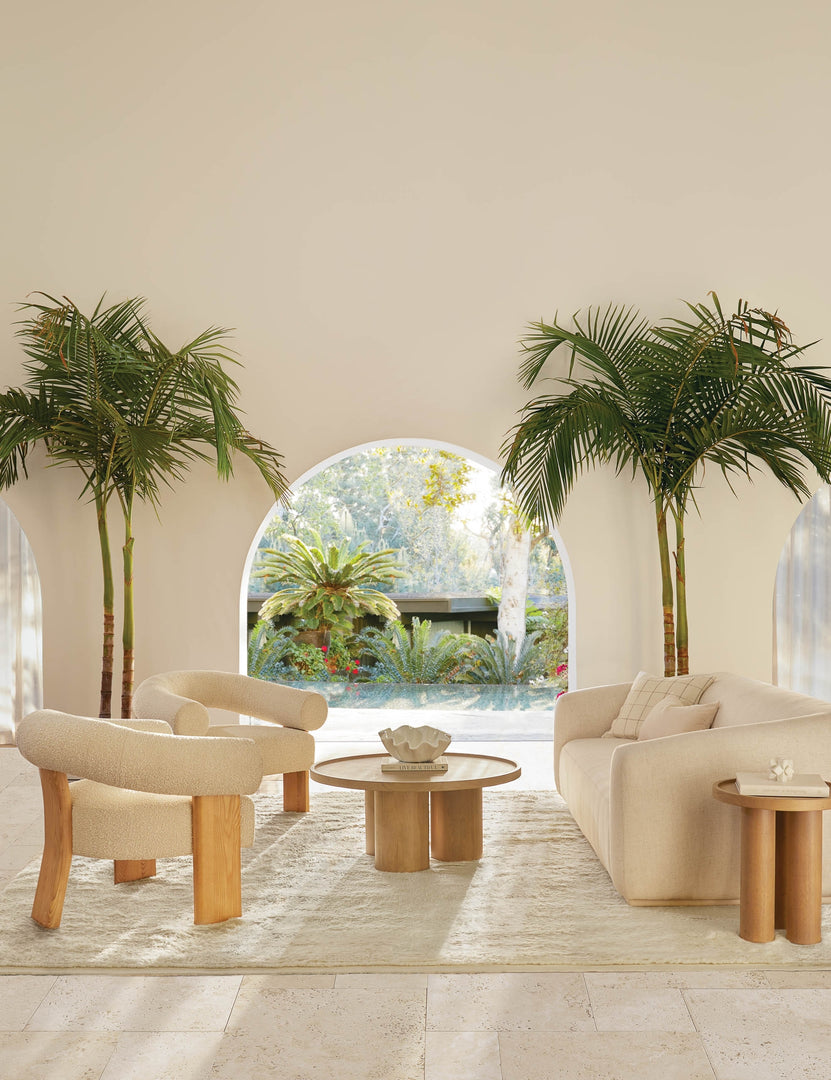 #color::cream | Two Celeste honey wood accent chairs with wishbone frames sit in a tropical living room with a white plush carpet and natural rounded sofa with a large archway in the background leading to the outdoors.