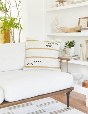 The Iconic ivory stripe square pillow sits on a white sofa with a wood frame with a decor filled shelf in the background