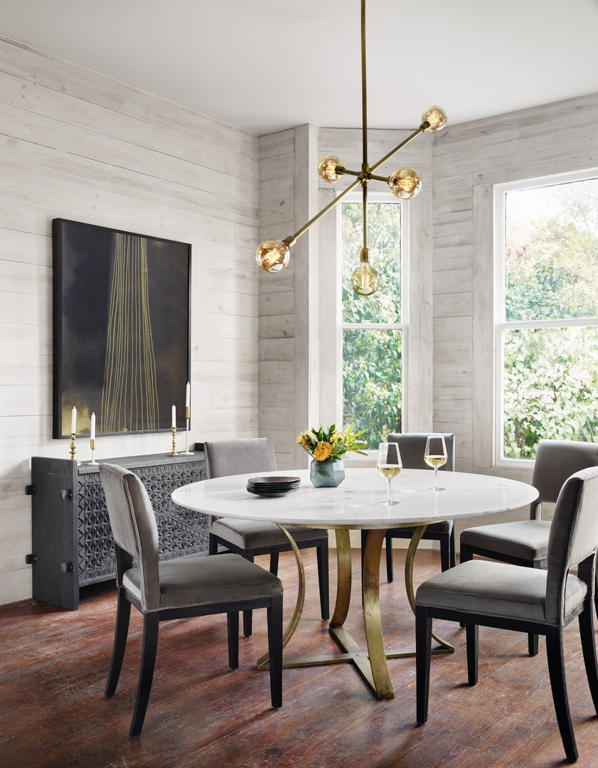 | The Brea round dining table with marble top and gold base sits in a dining room with gray velvet dining chairs, a gold and black wall art, and a sculptural chandelier 