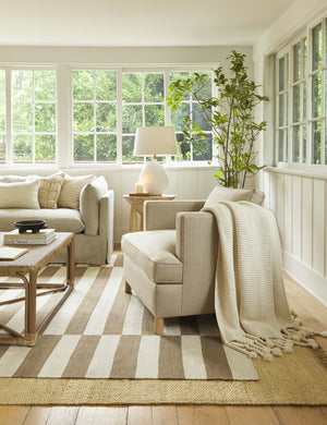 The Belmont stripe linen accent chair sits atop two layered rugs in a room with wrap-around windows and a woven coffee table