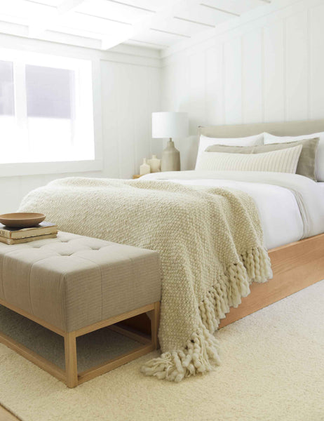 #color::ivory | The Olema ivory handwoven throw with fringed ends lays in a bedroom with a cream plush rug, a wooden-base bed, and a beige cushioned bench