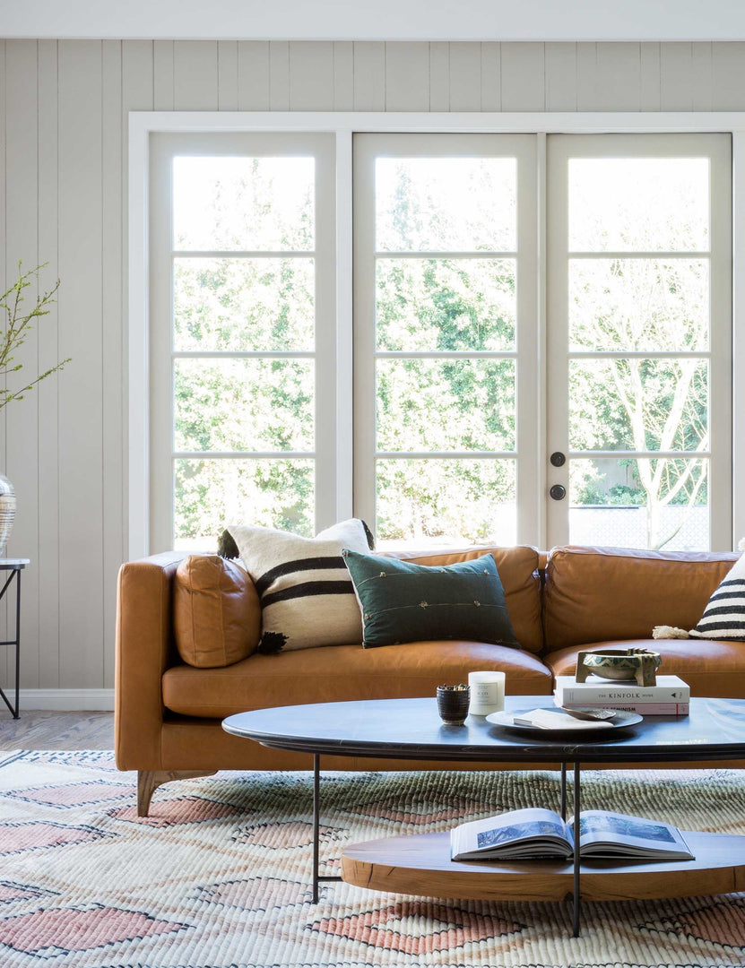 | The Jocelyn brown leather sofa sits in a living room in front of white-framed french doors, behind a black oval coffee table, sitting atop a diamond patterned rug.