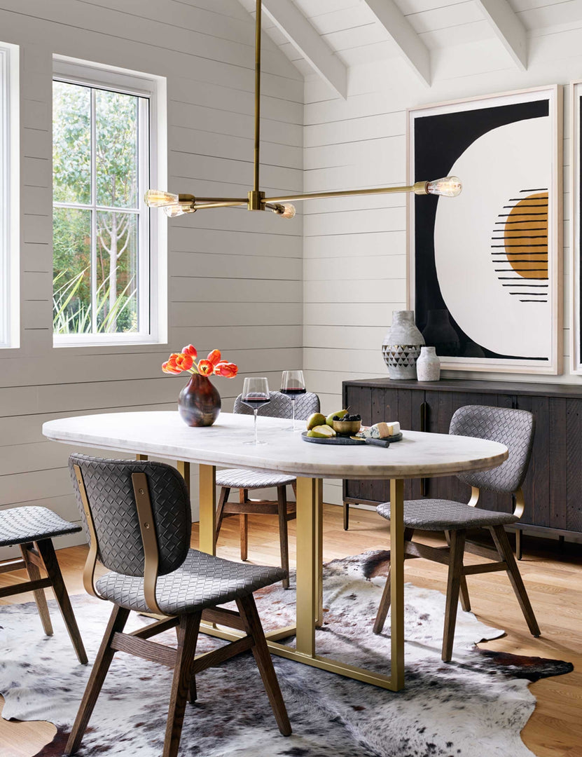 | The Kara oval dining table with white marble top and brass base sits in a dining room with an animal skin rug, black woven dining chairs, and white paneled walls