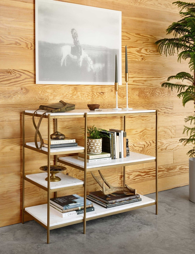 | The Kathleen console table with marble shelves and gold finish sits in a room with wooden paneled walls, a gray stone floor, with books and sculptural objects stacked within its shelving.