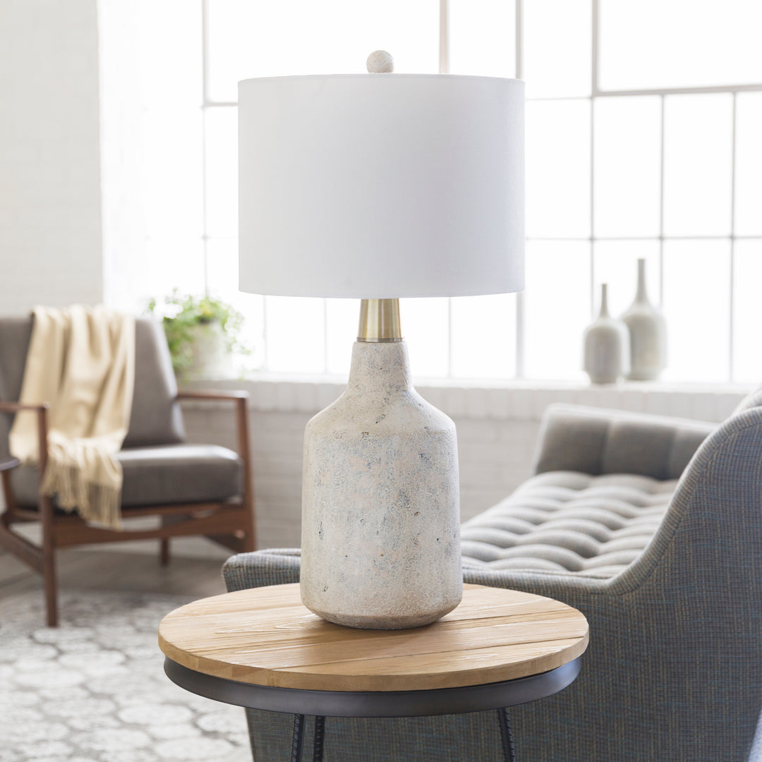 | The Langley table lamp with stone base and white finial sits in a living room atop a wooden side table with a gray accent chair and sofa in the background